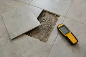 A lifted floor tile exposes grooved mortar beside a pinless moisture meter.