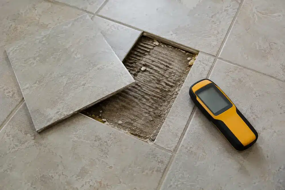 A lifted floor tile exposes grooved mortar beside a pinless moisture meter.